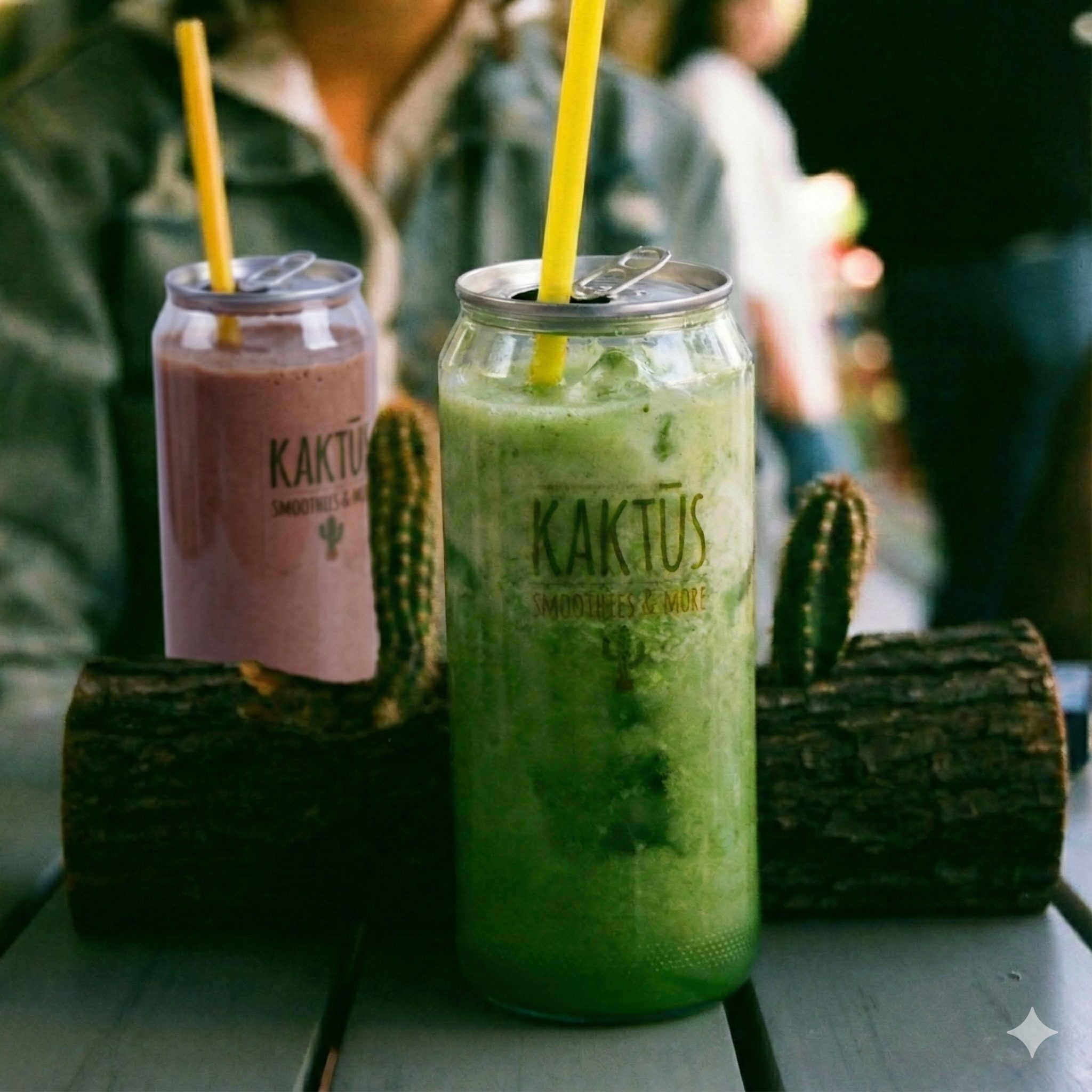 Two canned smoothies labeled "Kaktus" with straws, one pink and one green, on a table with cacti and wood logs in the background.